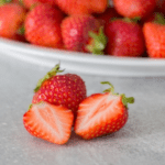 Im Vordergrund eine ganze und eine halbierte Erdbeere, im Hintergrund eine Schale mit vielen Erdbeeren